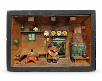 Op de afbeelding: Een houten dioramadoos met een uitgesneden scène van twee figuren die elkaar omhelzen in een rustieke kamer. Het interieur bevat een groene tegelkachel, een kleine tafel met een rode basis, een wandklok en planken met miniatuurvoorwerpen.