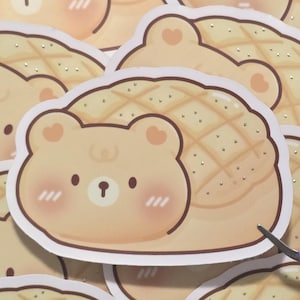 May include: A die-cut sticker featuring a light brown bear character with a rounded head, small ears, and pink cheeks. The bear is attached to a tan, grid-patterned bread loaf shape with small silver glitter accents scattered across the surface.
