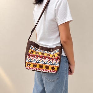 May include: A brown crossbody bag featuring a patterned panel with repeating brown bear faces, yellow backgrounds, and horizontal rows of red and blue geometric zig-zag and diamond shapes. The bag has a solid brown top border with two metal rivets.