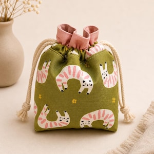 May include: A small olive green drawstring pouch featuring a repeating pattern of white cats with pink stripes and small yellow flowers. The bag has a soft pink interior lining and thick, cream-colored braided rope drawstrings on both sides.