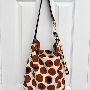 Puede incluir: Un bolso de hombro estilo hobo hecho de tela color crema con grandes lunares marrones. Algunos lunares contienen iconos negros de artículos relacionados con el café y la palabra coffee. El bolso tiene una correa larga marrón oscuro.
