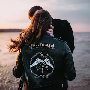 May include: A black leather jacket featuring a white graphic on the back. The design displays two skeleton hands touching fingers, a star, lightning bolts, and the text "TILL DEATH" above and "MMXXIII" below, all enclosed in a circular frame.