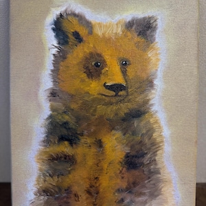 May include: An oil painting on canvas depicting a bear cub with textured, golden-brown fur and dark patches. The bear is centered against a light beige background with a soft white halo effect around its silhouette. The artist's initials SP are visible.