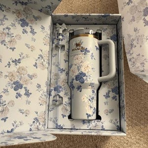 以下が含まれることがあります： 花柄のライトブルーのスタンレー製タンブラーと、リボン付きのストローがセットになった、お揃いの花柄ギフトボックス入り商品。タンブラーの前面にはスタンレーのロゴとLoveShackFancyの文字が印字されています。