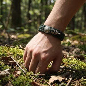 Pode incluir: Uma pulseira de paracord verde-escura com dois berloques metálicos: um escudo circular com superfície texturizada e um capacete em miniatura. A pulseira é feita de corda trançada e prende-se ao pulso com um mecanismo de fecho integrado.