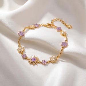 Könnte beinhalten: Ein goldfarbenes Kettenarmband mit abwechselnden blumenförmigen Anhängern und einem zentralen Sonnenstrahlen-Anhänger. Die Anhänger sind mit kleinen, klaren weißen Kristallen und hellvioletten Steinen besetzt. Mit Karabinerverschluss und Verlängerungskette.