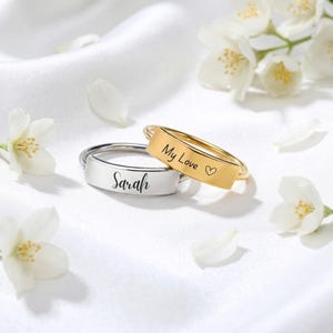 Pode incluir: Dois anéis de metal com placas retangulares planas. Um anel é prateado e gravado com o nome Sarah em letra cursiva. O outro anel é dourado e gravado com o texto My Love seguido por um pequeno símbolo de coração.