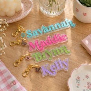 Pode incluir: Quatro porta-chaves de acrílico com fechos lagosta dourados, cada um com um nome numa cor diferente: Savannah em azul claro, Maddie em rosa choque, Brynn em verde lima e Kelly em lavanda. Cada porta-chaves inclui um pequeno pendente.