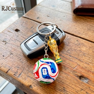 May include: A metal keychain featuring a miniature gold-colored trophy charm and a spherical soccer ball charm with white, red, blue, and green geometric patterns, attached to a silver-toned split ring resting on a black car key fob.