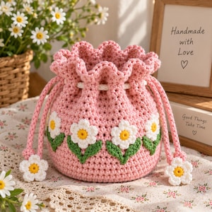May include: A light pink crochet drawstring pouch featuring a repeating pattern of white daisies with yellow centers and green leaves. The bag has a ruffled top edge, a white drawstring cord, and two dangling white daisy charms at the ends.