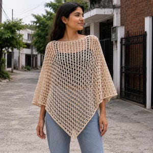 May include: A beige crochet poncho featuring an openwork diamond pattern with a wide boat neckline and a pointed hemline, worn over a black top and light blue denim jeans. The garment has a loose, airy construction with short, wide sleeves.