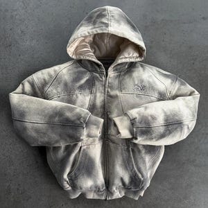May include: A zip-up hooded jacket featuring a distressed, washed-out grey and off-white finish. The garment includes a quilted cream-colored lining inside the hood, ribbed cuffs and hem, and a small embroidered logo on the upper left chest.