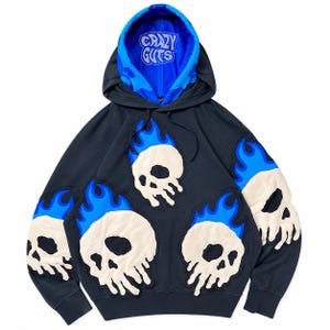 May include: A black hooded sweatshirt featuring multiple off-white skull appliques with bright blue flame accents. The interior of the hood is bright blue and displays the text "CRAZY GUTS" in a stylized white font. The sleeves and front feature skulls.