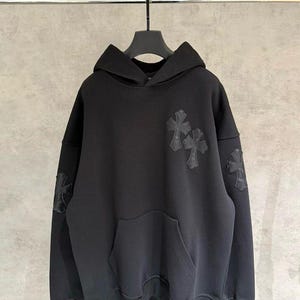 May include: A black long-sleeve hooded sweatshirt featuring a large hood, a front kangaroo pocket, and ribbed cuffs and hem. The garment is decorated with three black cross-shaped patches, with two positioned on the chest and one on the sleeve.