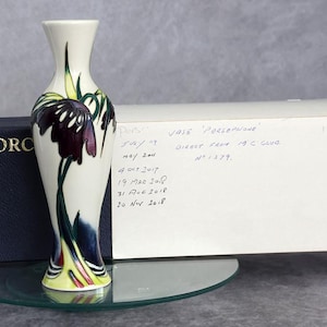Peut inclure: Un vase en céramique de couleur crème orné d'un motif floral peint à la main avec des accents violet foncé, vert et jaune. Le vase est placé devant une boîte bleu foncé marquée MOORC et une boîte blanche avec du texte manuscrit VASE PERSEPHONE.
