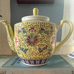 May include: A yellow ceramic teapot featuring a repeating black scroll pattern, floral motifs in pink and green, and red Chinese characters within circular frames. The teapot has a matching lid with a small knob, a curved handle, and a spout.