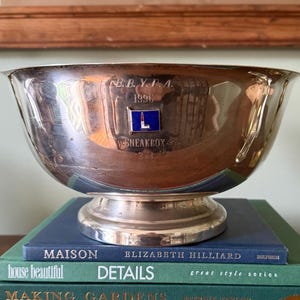 May include: A silver-toned metal trophy bowl featuring an engraved inscription that reads B.B.Y.R.A. 1996 SNEAKBOX 2nd. A small rectangular blue enamel emblem with a white lighthouse graphic is centered on the front of the polished bowl.