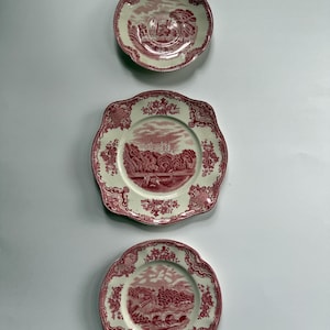 May include: Three ceramic plates featuring a red and white transferware pattern. Each plate displays a detailed landscape scene in the center, surrounded by an intricate floral border. The plates have scalloped edges and a cream-colored base.