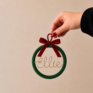 May include: A circular ornament featuring a dark green braided rope frame and a deep red velvet ribbon bow at the top. The centre is a clear disc with the name Ellie written in a thin, gold-coloured cursive wire script across the middle.