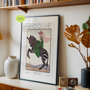 May include: A framed wall art print featuring a vintage Vogue magazine cover illustration. The artwork depicts a person in a green coat and pink hat riding a rearing zebra, with two giraffes in the background. The text VOGUE is printed at the top.
