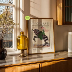 May include: A framed Vogue magazine print featuring an illustration of a person riding a zebra, displayed next to a dark brown vase with dried flowers and a tall amber glass vase. The print includes the text VOGUE and a small green DIGITAL label.