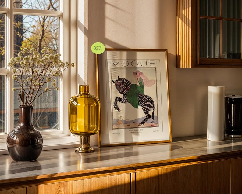 May include: A framed Vogue magazine print featuring an illustration of a person riding a zebra, displayed next to a dark brown vase with dried flowers and a tall amber glass vase. The print includes the text VOGUE and a small green DIGITAL label.