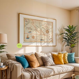 Puede incluir: Una impresión enmarcada de un mapa antiguo cuelga sobre un sofá seccional beige con cojines coloridos. Una mesa auxiliar de madera con una lámpara y una planta en maceta están en primer plano. La habitación tiene una paleta de colores neutros.