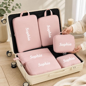 Peut inclure: Un ensemble de six cubes de rangement rose clair disposés à l'intérieur d'une valise ouverte couleur crème. Chaque organisateur en tissu rectangulaire possède une poignée, une fermeture éclair et le prénom Sophia imprimé en blanc.