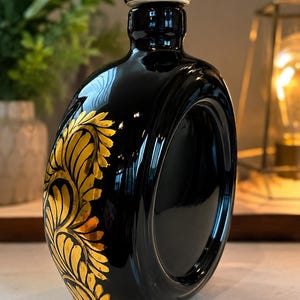 Puede incluir: Un decantador de cerámica negra brillante con un centro circular hueco y un tapón a juego. El exterior presenta un patrón decorativo de pan de oro con hojas estilizadas y enredaderas en espiral sobre el lado curvo del recipiente.