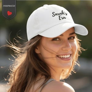 Puede incluir: Una gorra de béisbol blanca con el texto bordado en negro Sarah's Era en el panel frontal. La gorra tiene una corona estructurada, una visera curva y un botón en la parte superior. Se observa una pequeña etiqueta gris oscuro con la palabra PERSONALIZE y un corazón rojo.
