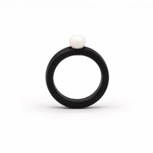 May include: A circular ring featuring a matte black band with a smooth, round white bead centered at the top. The band has a consistent thickness and a dark, textured finish, contrasting with the bright, spherical white accent piece.