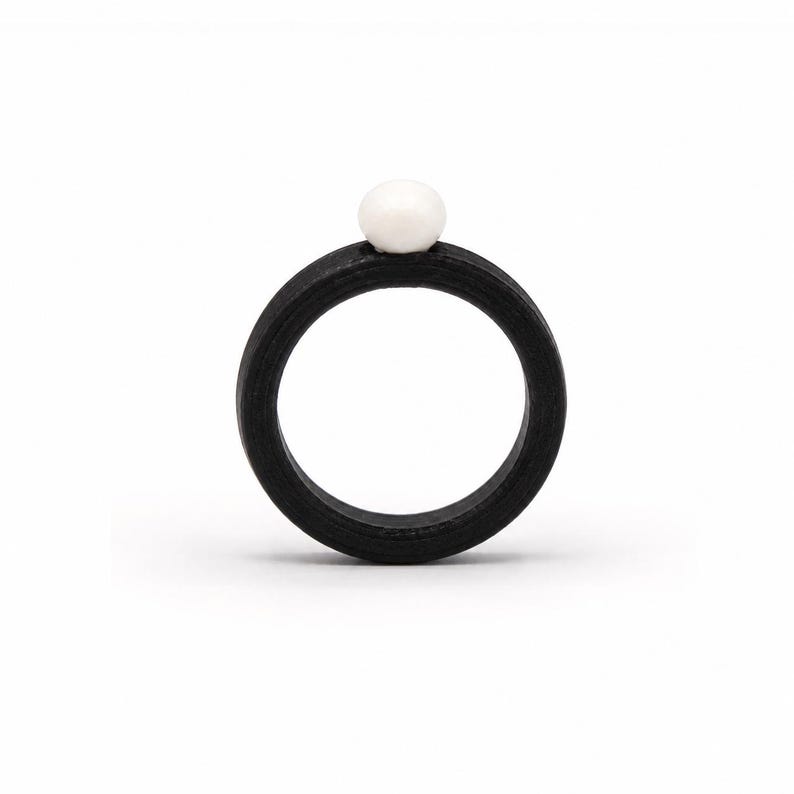 May include: A circular ring featuring a matte black band with a smooth, round white bead centred at the top. The band has a consistent thickness and a dark, textured finish, contrasting with the bright, spherical white accent piece.