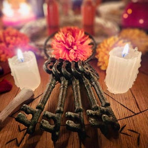 May include: A cluster of four dark, weathered metal skeleton keys arranged in a fan shape, topped with a vibrant orange flower. The keys feature ornate bows and notched bits, resting on a wooden surface marked with etched geometric lines.