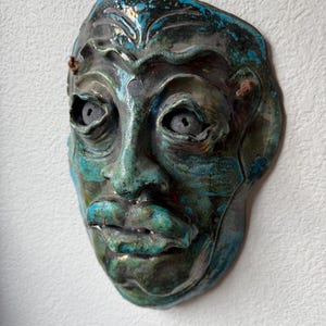 May include: A ceramic wall mask featuring a glossy, mottled glaze in shades of deep teal, turquoise, and dark brown. The mask displays a human-like face with prominent brow ridges, hollowed eye sockets, a defined nose, and full lips.