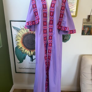May include: A floor-length lavender robe featuring wide, flowing sleeves and an open front. The garment is accented with a vibrant, geometric patterned trim in shades of red, pink, and dark blue along the neckline, sleeve edges, and hemline.