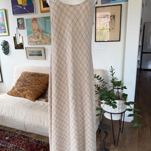 May include: A sleeveless, floor-length dress in a light beige color featuring a dark, thin, diagonal grid pattern. The garment has a wide, rounded scoop neckline and a straight, flowing silhouette, displayed on a dress form mannequin.