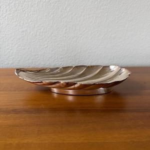 May include: A silver-toned metal dish shaped like a seashell with a scalloped edge and a smooth, curved interior. The dish features a raised, rounded base that elevates the shell-like form, creating a shallow, open container with a polished finish.
