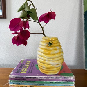 May include: A yellow and white ceramic beehive-shaped vase with a small bee accent, holding a branch of bright pink bougainvillea flowers, resting on a stack of seven colorful children's books with visible titles including Me Too Iguana.