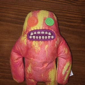 May include: A plush monster toy featuring a tie-dye pattern of vibrant pink and yellow. The creature has a large purple stitched mouth filled with white plastic teeth, an orange button for one eye, and a bright green button for the other eye.