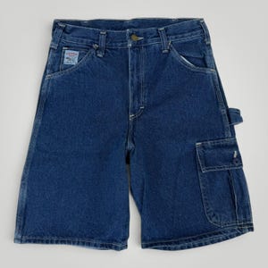 May include: Dark blue denim shorts featuring a classic five-pocket design with a button fly, belt loops, and a utility pocket on the right leg. A rectangular white label with blue text and a dog graphic is stitched onto the front right pocket area.