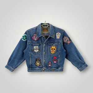 May include: A blue denim jacket featuring a button-up front, two chest pockets, and multiple embroidered patches including colorful sugar skulls, a Wyoming emblem, and a circular green patch on the sleeve. The jacket has a classic collar and metal buttons.