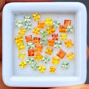 May include: A collection of small, square-cut gemstones in a white display box. The stones feature a vibrant color palette of yellow, orange, and light green, each with a faceted surface that reflects light, arranged loosely on a white foam insert.