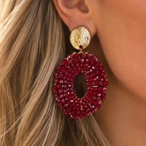 Può includere: Un orecchino color oro caratterizzato da un perno circolare testurizzato collegato a un grande elemento pendente di forma ovale. Il pendente è densamente ricoperto di piccole perline sfaccettate rosso bordeaux scuro in cerchio.
