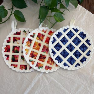 May include: Three round fabric pot holders designed to look like fruit pies. Each features a printed pattern of red apples, peaches, or blueberries, overlaid with a white lattice crust design, a scalloped edge, and a small fabric hanging loop.