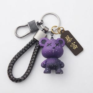 以下が含まれることがあります： 紫色のクマのフィギュアが付いたキーホルダー。白い斑点模様とX型の目が特徴です。ダークグレーの編み込みコード、銀色の金属製クラスプ、二重リング、そして「暴富」という金色の漢字が刻まれたダークブラウンの長方形タグが付いています。