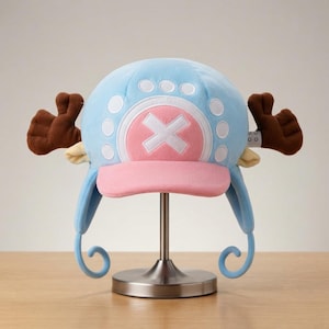 Puede incluir: Un gorro de felpa azul claro con visera rosa y un círculo rosa central con un símbolo X blanco. El gorro incluye astas de felpa marrón a cada lado, pequeñas orejas de color crema y dos largas solapas laterales azules con extremos rizados.