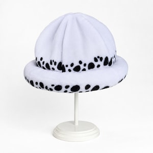 May include: A white plush bucket hat featuring a black spotted pattern around the brim and the base of the crown. The hat has a soft, fuzzy texture and a rounded top with visible seams, displayed on a simple white stand against a plain background.