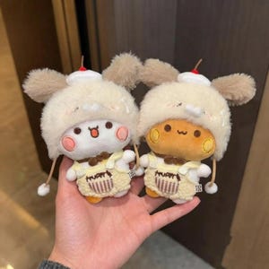 May include: Two small plush toys featuring round heads with pink cheeks, wearing fluffy cream-colored hats with brown ears and a small red cherry on top. Each toy wears yellow overalls with a small pocket and brown bow tie detail at the neck.
