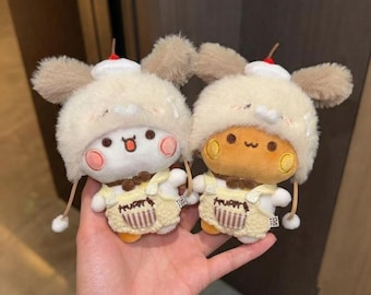 Può includere: Due piccoli peluche con teste rotonde e guance rosa, che indossano cappelli di pelliccia color crema con orecchie marroni e una piccola ciliegia rossa. Ogni peluche indossa una salopette gialla con una tasca e un papillon marrone al collo.