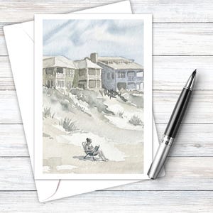 Puede incluir: Una tarjeta de felicitación en acuarela que muestra una escena de playa con casas en dunas y una persona sentada en una silla leyendo un libro. La tarjeta está junto a un bolígrafo negro y plateado. La obra usa tonos suaves azules, grises y beige.
